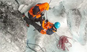Dwóch alpinistów w pomarańczowych kurtkach wspina się po lodowcu, używając lin i raków.