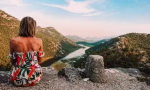 Kobieta w kwiecistej sukience podziwia widok na góry i rzekę. To inspiracja, jak zorganizować logistykę wyjazdu w góry bez własnego samochodu.