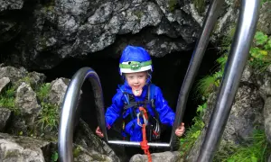 Dziecko w kasku i niebieskiej kurtce zjeżdża po poręczy w skalnym przejściu.