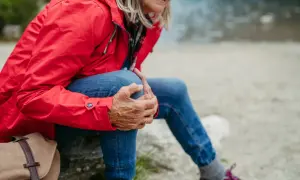 Kobieta w czerwonej kurtce i jeansach odpoczywa, masując kolano. To ważny moment, by pomyśleć, jak przygotować stawy kolanowe do długich zejść ze szczytów.