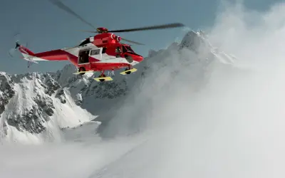 Czerwono-biały helikopter ratunkowy w akcji nad zaśnieżonymi górami. Sprawdź, jakie ubezpieczenie w góry za granicę obejmuje akcję z helikopterem.