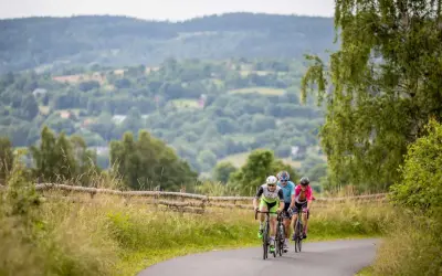 Trzech rowerzystów jedzie po asfaltowej drodze wśród zielonych wzgórz.