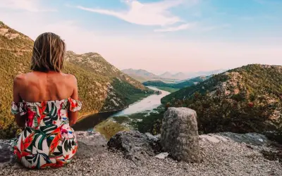 Kobieta w kwiecistej sukience podziwia widok na góry i rzekę. To inspiracja, jak zorganizować logistykę wyjazdu w góry bez własnego samochodu.