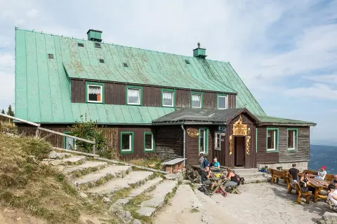 Schronisko pod Łabskim Szczytem, drewniany budynek z zielonym dachem, gdzie turyści odpoczywają przy stolikach.