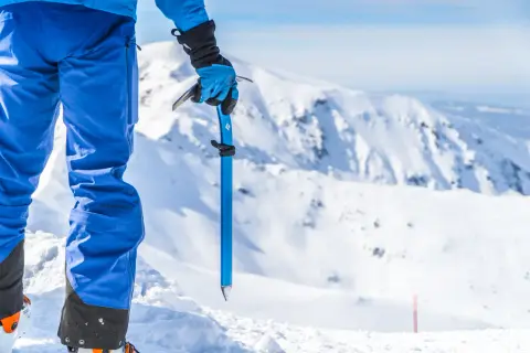 Osoba w niebieskich spodniach i rękawicach trzyma czekana, gotowa do wspinaczki. Pokazuje, jak prawidłowo używać czekana i raków na stromym lodzie.