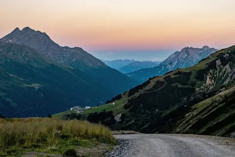 Jak zaplanować trasę samochodową przez przełęcze alpejskie widoki? Droga wije się przez zielone zbocza, w oddali widać ośnieżone szczyty i domki.