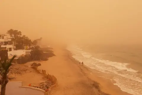 Pomarańczowe niebo i plaża na Wyspach Kanaryjskich, widoczne skutki silnego wiatru calima na szlakach.