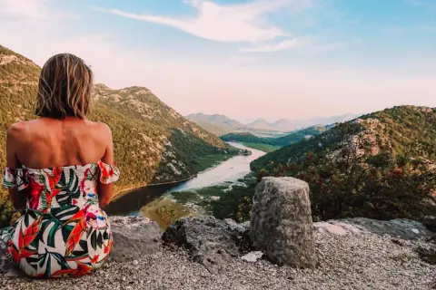 Kobieta w kwiecistej sukience podziwia widok na góry i rzekę. To inspiracja, jak zorganizować logistykę wyjazdu w góry bez własnego samochodu.