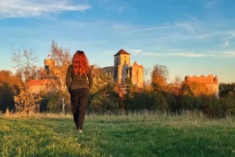 Kobieta z rudymi włosami idzie w stronę ruin zamku. To krajobraz inny niż krakowskie kopce, ale równie piękny jak trasy w Jurze Krakowsko-Częstochowskiej.