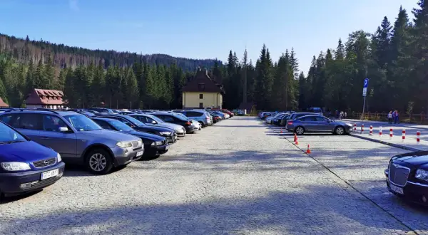 Duży parking przy Palenicy Białczańskiej, pełen samochodów, z górami i lasem w tle.