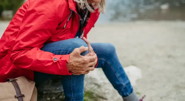 Kobieta w czerwonej kurtce i jeansach odpoczywa, masując kolano. To ważny moment, by pomyśleć, jak przygotować stawy kolanowe do długich zejść ze szczytów.