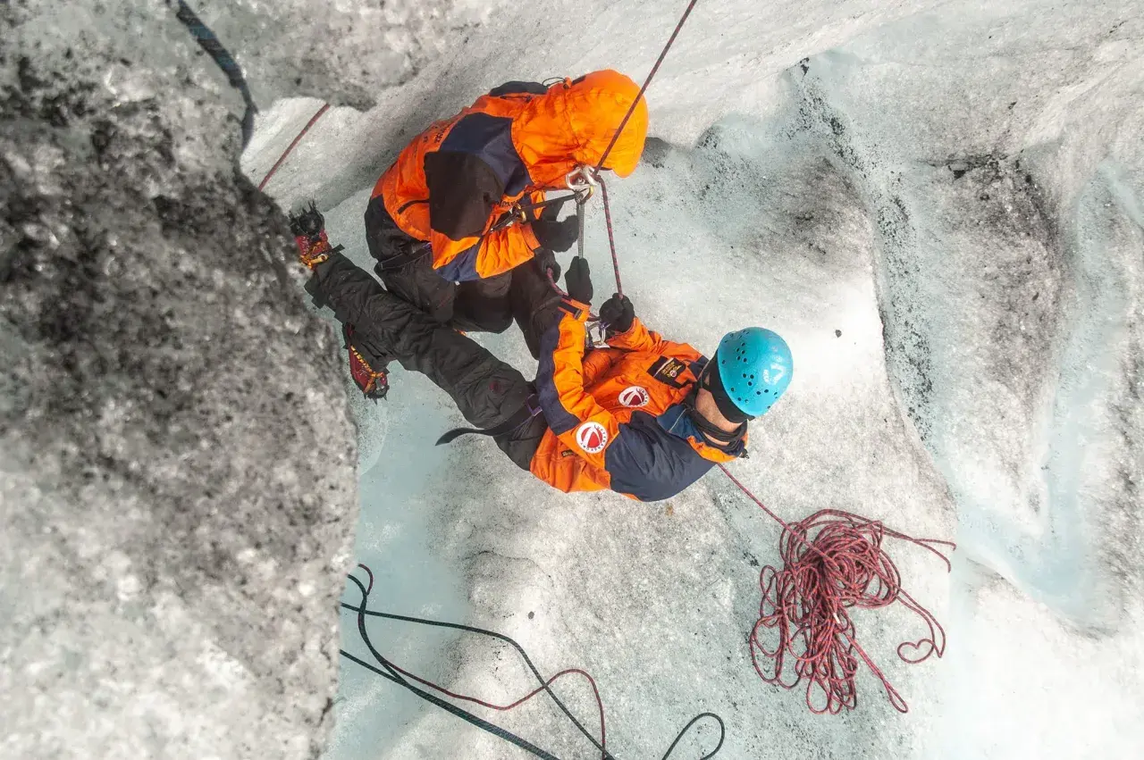 Dwóch alpinistów w pomarańczowych kurtkach wspina się po lodowcu, używając lin i raków.