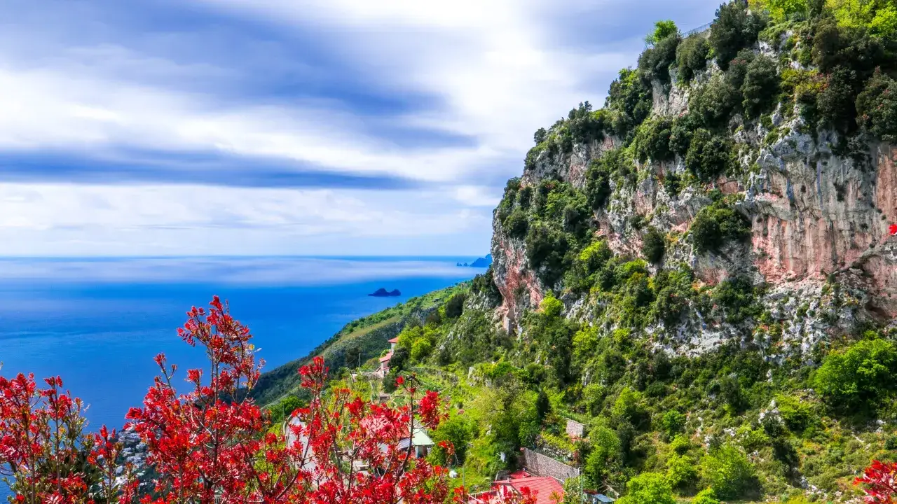 Widok na wybrzeże Amalfi i wyspę Capri. Krwistoczerwone liście na pierwszym planie, skaliste zbocze porośnięte zielenią, błękitne morze i chmury.