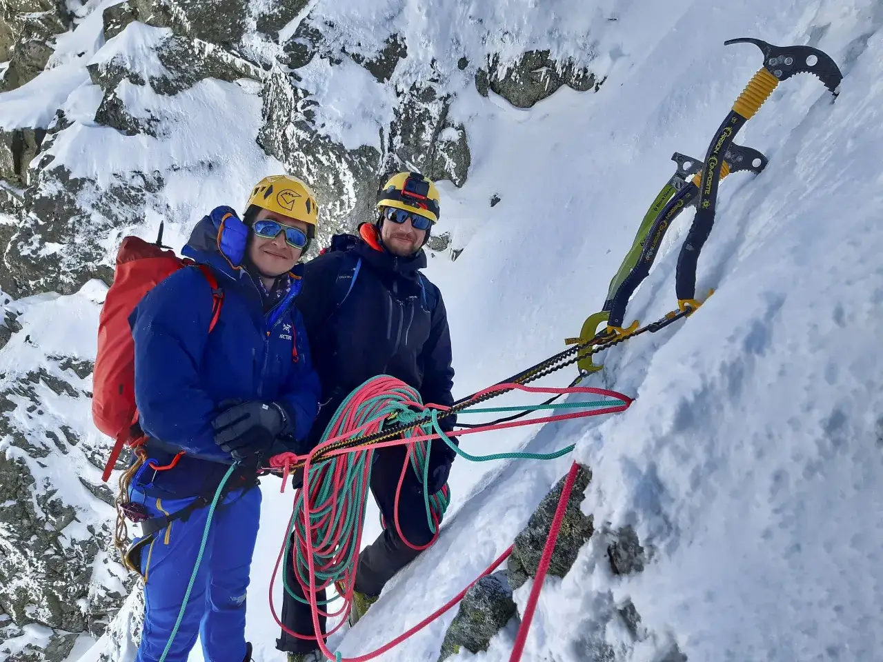 Dwaj alpiniści wspinają się po ośnieżonym zboczu, gotowi do asekuracji. Widać sprzęt wspinaczkowy, liny i czekany.