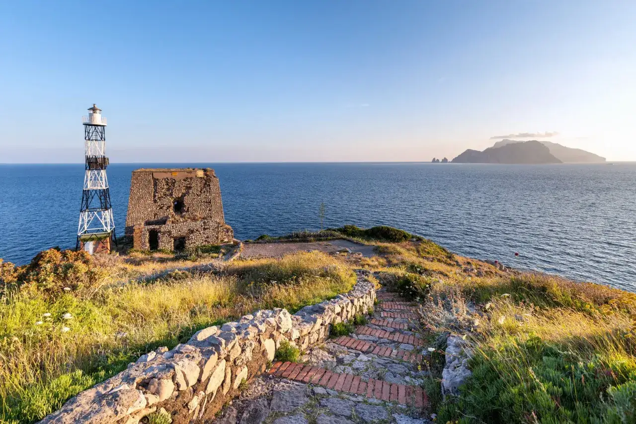 Kamienna ścieżka prowadzi do latarni morskiej i ruin na klifie. W oddali widać wyspę Capri, a morze otacza wybrzeże Amalfi.
