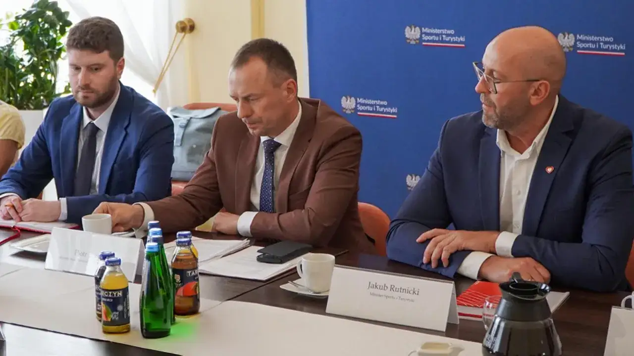 Trzech mężczyzn przy stole, dyskusja o tym, jak dobrać tempo marszu do najsłabszego uczestnika wyprawy. Na stole napoje i dokumenty.