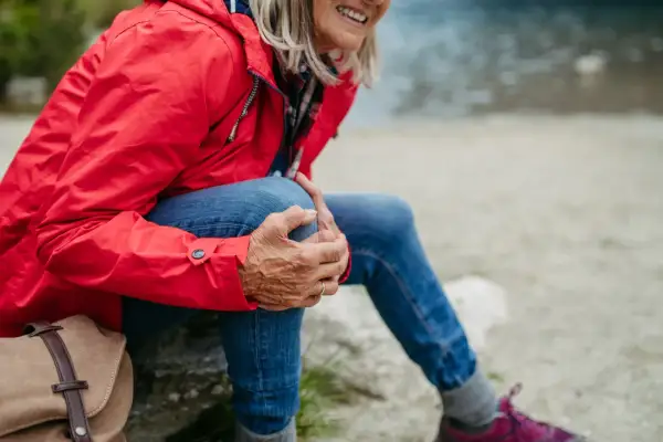 Kobieta w czerwonej kurtce i jeansach odpoczywa, masując kolano. To ważny moment, by pomyśleć, jak przygotować stawy kolanowe do długich zejść ze szczytów.