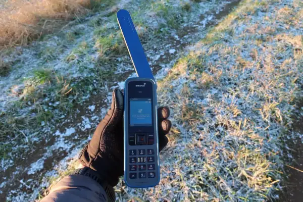 Ręka w rękawiczce trzyma telefon satelitarny Inmarsat IsatPhone, pokazując ekran z informacją o wyszukiwaniu sygnału. Idealne narzędzie, jak sprawdzić prognozę pogody przez telefon satelitarny na wyprawie.